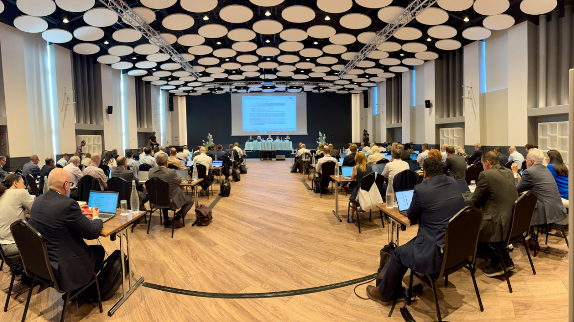 Konferenču zāle ar dalībniekiem pie galdiem, kuri klausās prezentāciju un diskusiju uz skatuves.