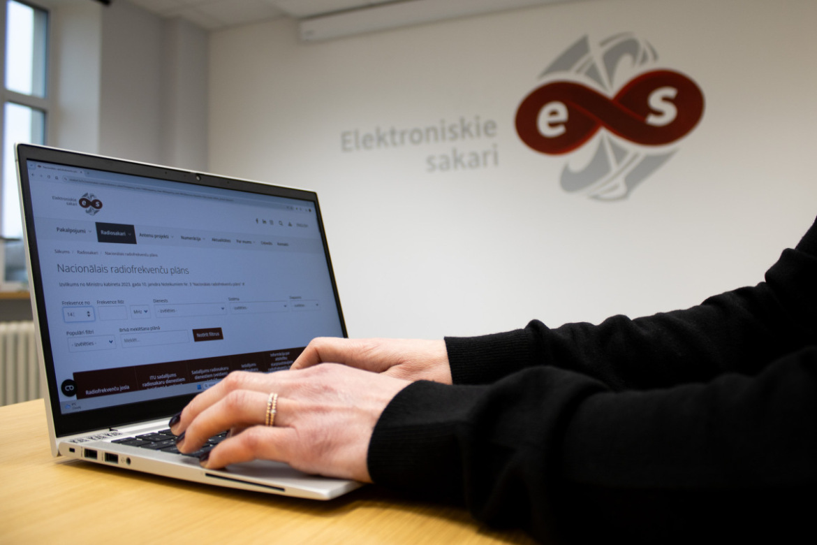 Persona izmanto datoru ar atvērtu Nacionālā radiofrekvenču plāna digitālo pārlūku, fonā redzams SIA "Elektroniskie sakari" logo