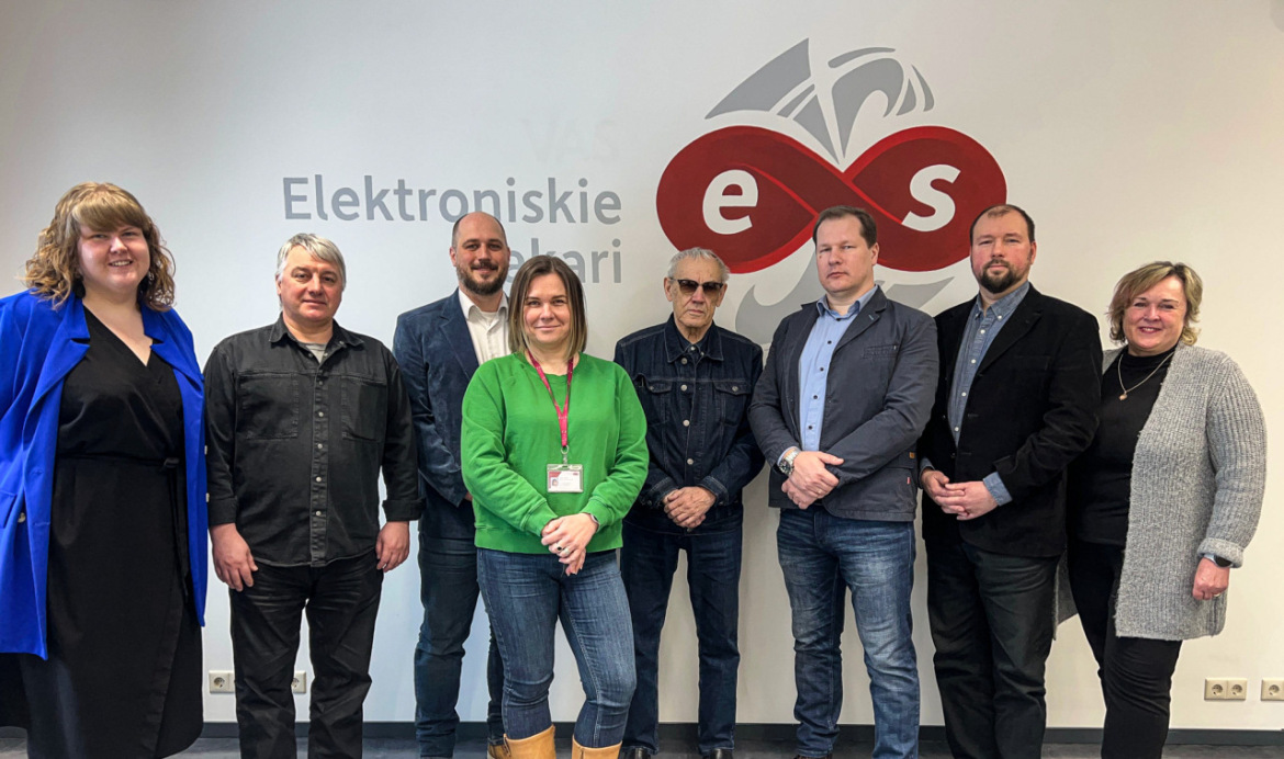 Astoņi cilvēki stāv pie sienas ar SIA “Elektroniskie sakari” logotipu