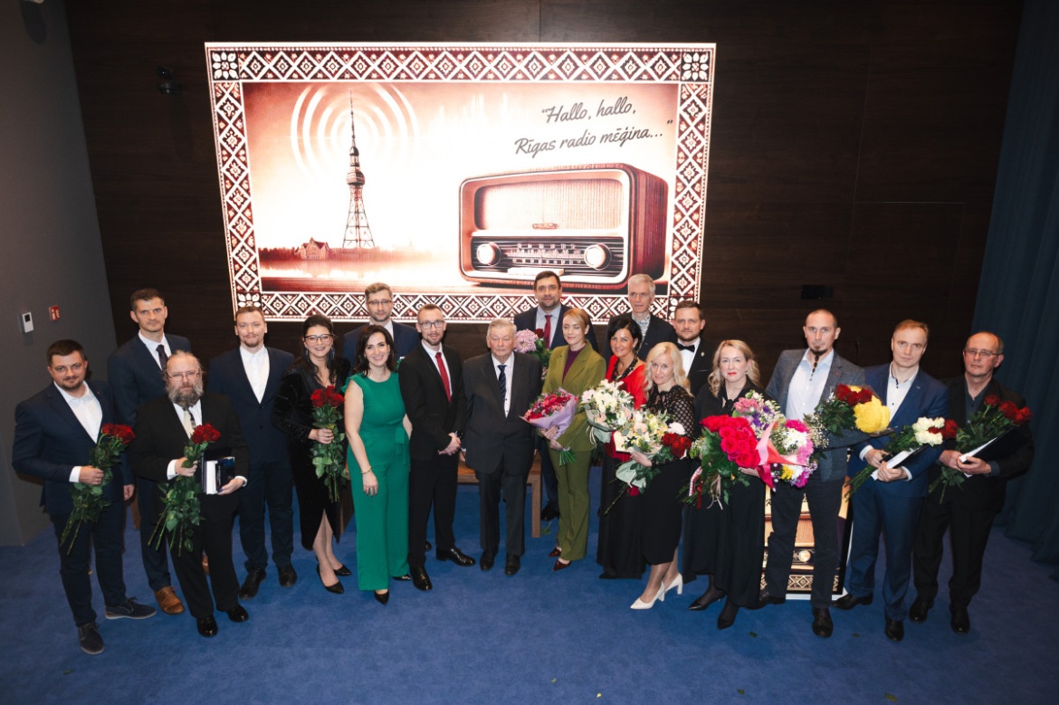 Grupas foto ar cilvēkiem - balvas laureātiem - svinīgā pasākumā; fonā attēlots Rīgas radio tornis un radio aparāts ar uzrakstu “Hallo, hallo, Rīgas radio mēģina…