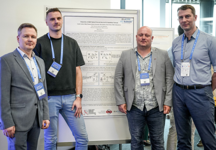 Četri vīrieši stāv posteru sesijā pie zinātniskā plakāta ar nosaukumu 'Integration of QKD Optical Network Into the 5G Standalone Network' Eiropas konferencē 'EuCNC & 6G Summit'. Visi valkā dalībnieku identifikācijas kartes un pozē kopbildē, fonā redzama izstādes vide ar citiem plakātiem un cilvēkiem.