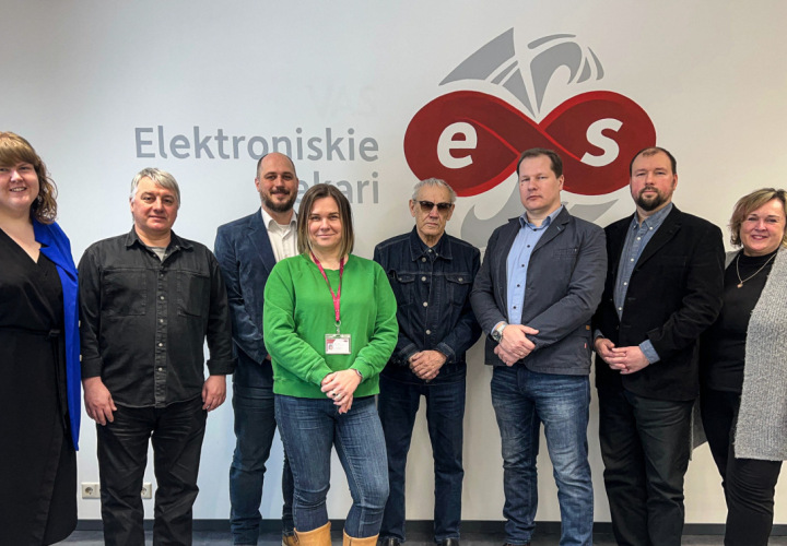 Astoņi cilvēki stāv pie sienas ar SIA “Elektroniskie sakari” logotipu