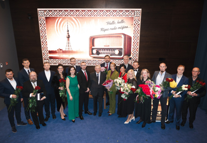 Grupas foto ar cilvēkiem - balvas laureātiem - svinīgā pasākumā; fonā attēlots Rīgas radio tornis un radio aparāts ar uzrakstu “Hallo, hallo, Rīgas radio mēģina…