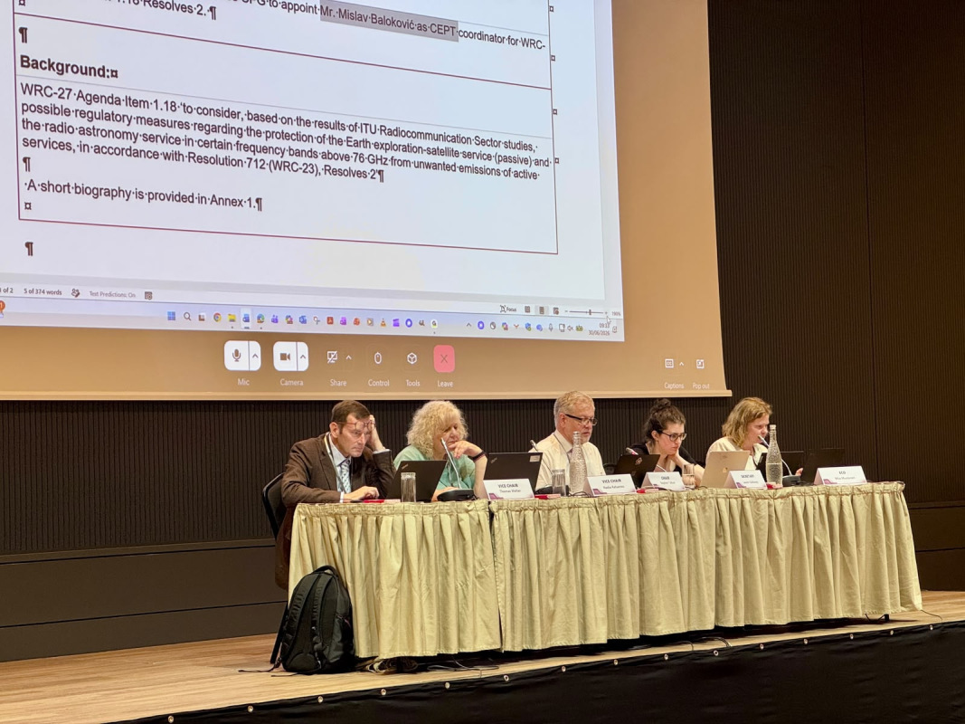 Pieci cilvēki sēž pie paneļdiskusijas galda konferencē, fonā uz ekrāna redzams prezentācijas dokuments