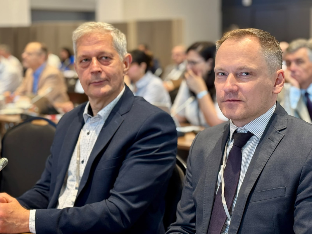 Divi vīrieši uzvalkos sēž konferencē kopā ar citiem dalībniekiem un skatās kameras virzienā.