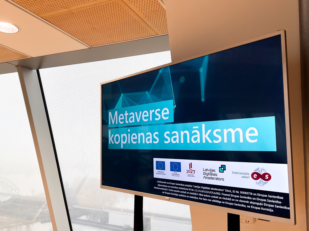 Attēlā redzams digitāls ekrāns ar uzrakstu “Metaverse kopienas sanāksme” un informāciju par projekta atbalstītājiem. Ekrānā redzami dažādi logotipi, tostarp Eiropas Savienības, Latvijas Digitālās Akseleratora un SIA “Elektroniskie sakari” logotipi. Fonā caur logiem redzama miglaina ainava ar tiltu un upi.