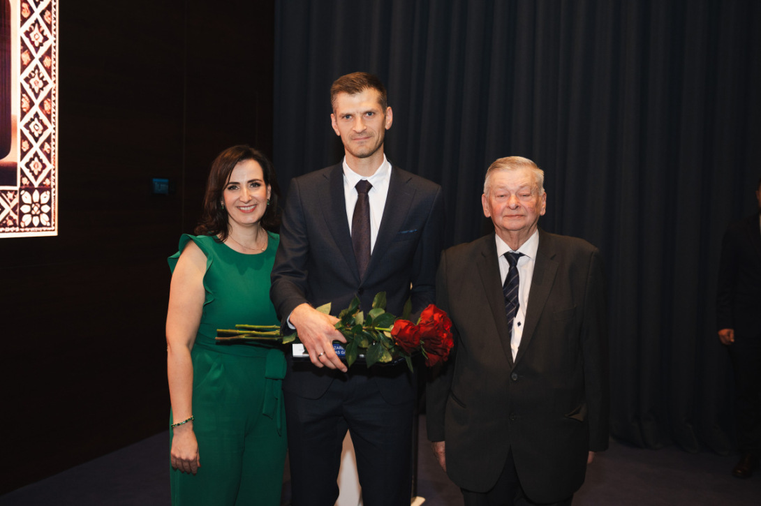 Trīs cilvēki svinīgā pasākumā, vidējais, laureāts, tur ziedus un balvu.