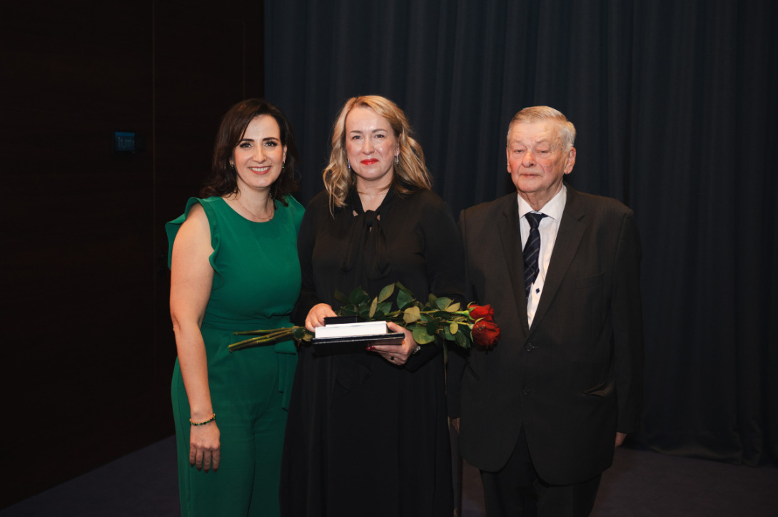 Trīs cilvēki svinīgā pasākumā, vidējā, laureāte, tur ziedus un balvu.